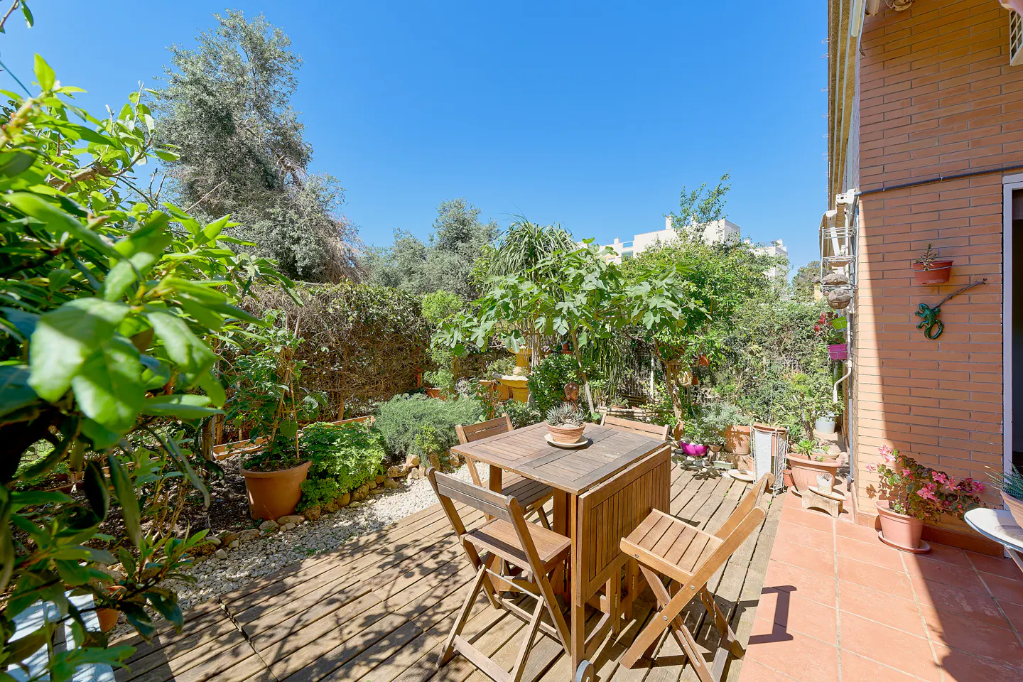 Outdoor patio with wooden table and chairs, surrounded by lush greenery and a brick building under a clear blue sky.