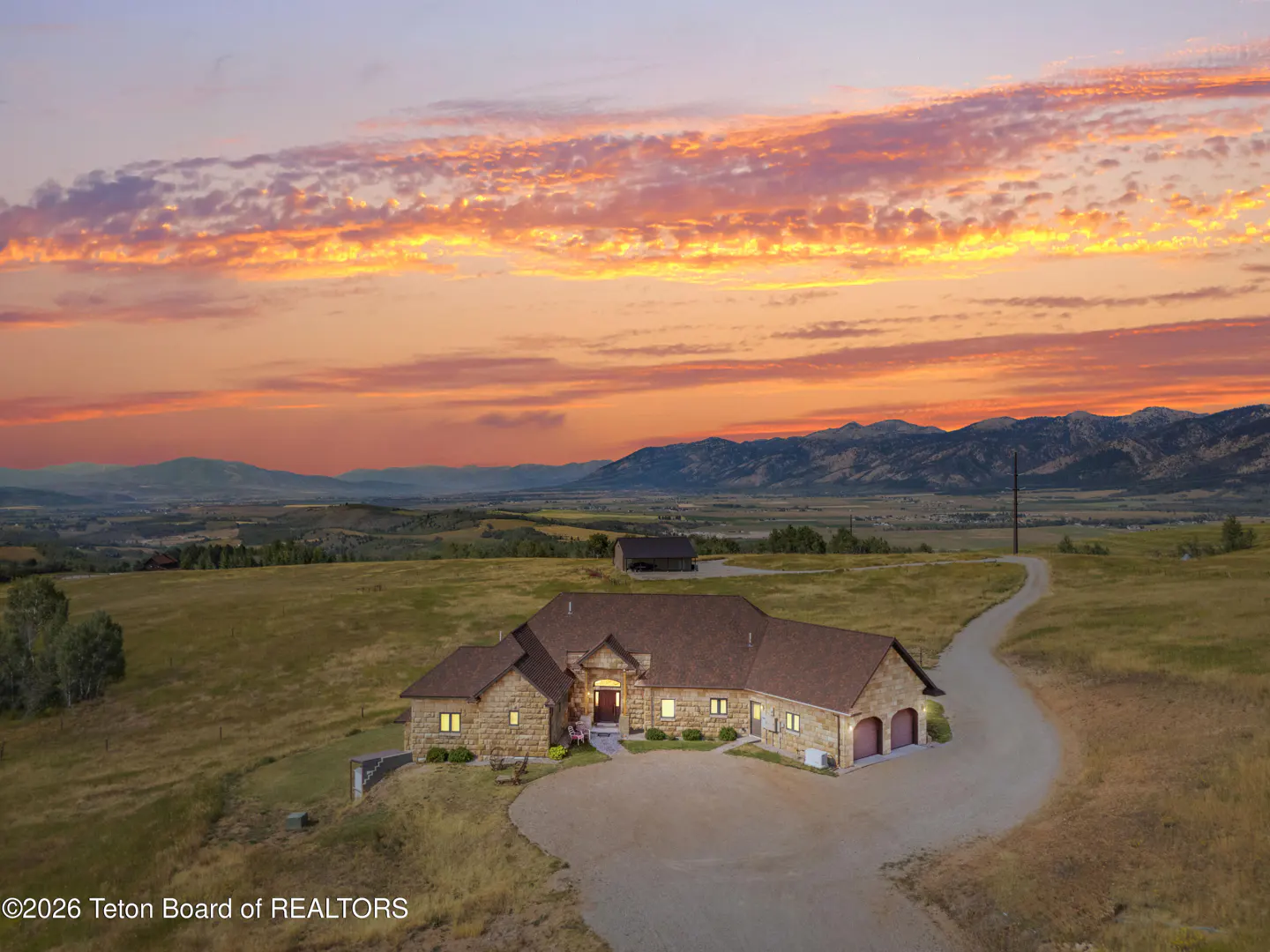A tan stone house with a brown roof sits on a hill under a colorful sunset sky. Mountains are in the background.
