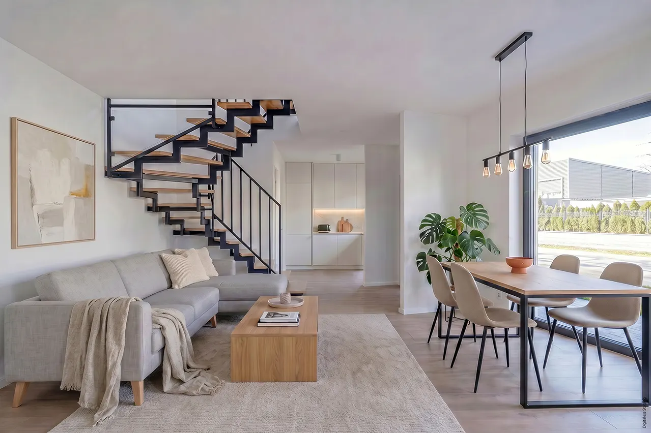 Bright, modern living space with a gray sofa, wooden stairs with black railings, and a dining table near a large window. A neutral rug covers the floor.