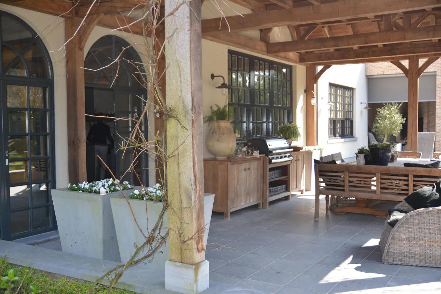 Outdoor patio with gray tile, wooden pergola, and furniture. Grill, cabinets, and planters add to the space.