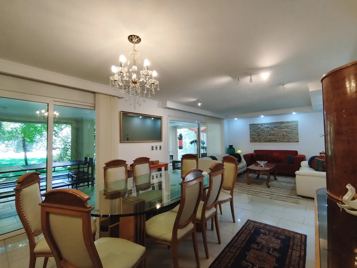 A bright, open-concept living and dining area with a glass table, wooden chairs, and a crystal chandelier. A red sofa and artwork are visible in the background.