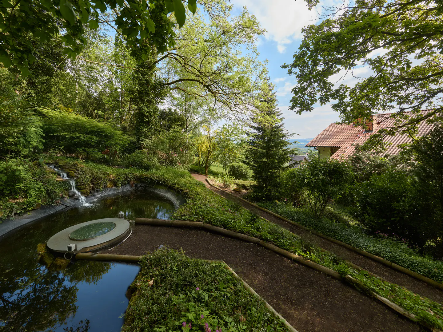 Lush garden with a pond, waterfall, and winding path leading to a house with a red tile roof. Green trees and blue sky.