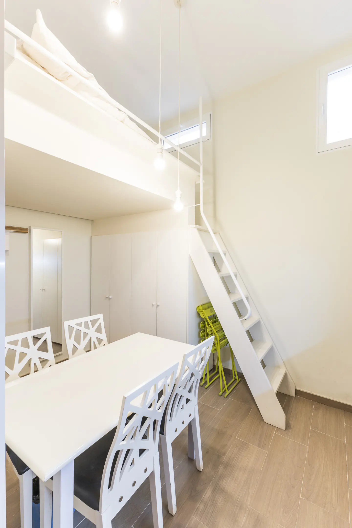 Bright room with a white table, chairs, and stairs leading to a loft bed. Green folding chairs are stacked near the stairs.