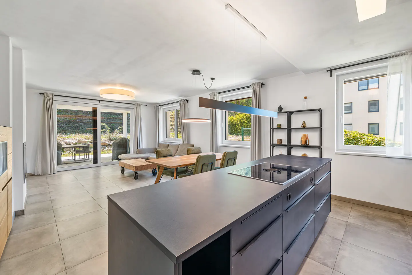 Bright, open-plan living space with a dark grey kitchen island, dining table, and a grey sofa. Sliding glass doors lead to an outdoor patio.