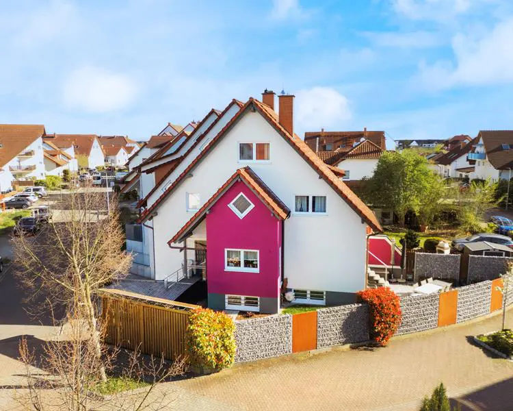 Two-story white house with a pink section and red roof, surrounded by a stone and wood fence.