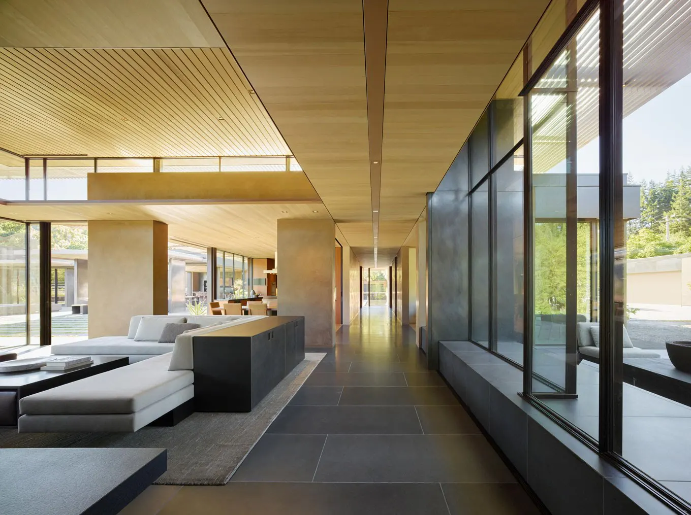 Interior view of a modern home with gray tile floors, wood ceilings, and large windows. A gray sofa and black cabinet are visible.