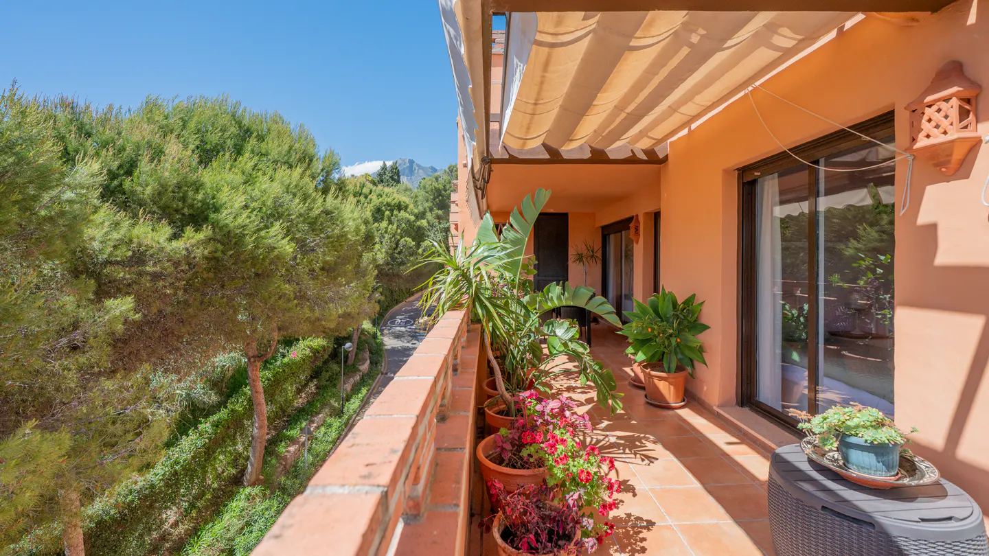 A sunlit balcony with potted plants, terracotta tiles, and a brick railing overlooks lush greenery and a distant mountain.