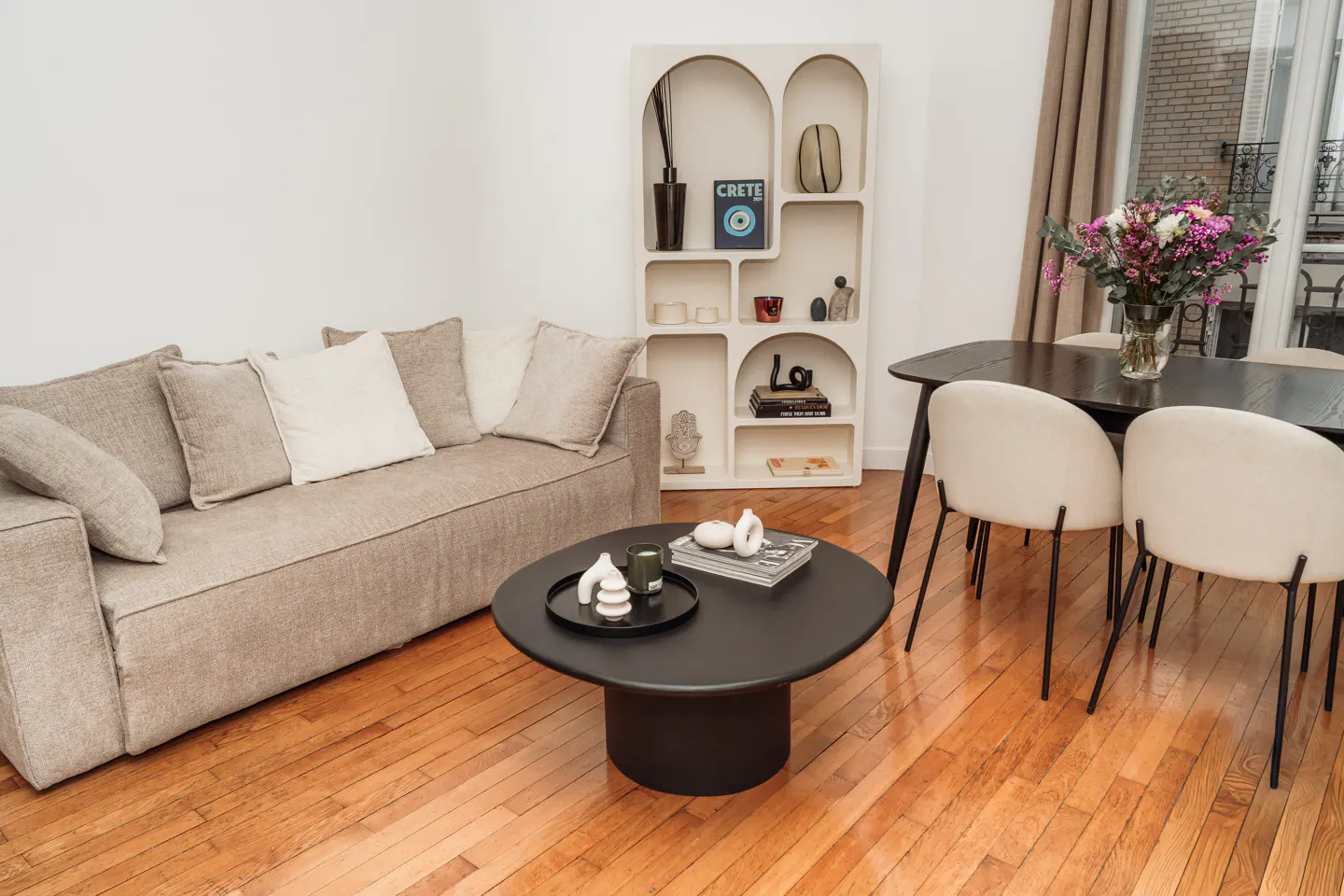 Living room with a beige sofa, black coffee table, and a white bookshelf with arched openings. Dining table with flowers near a window.