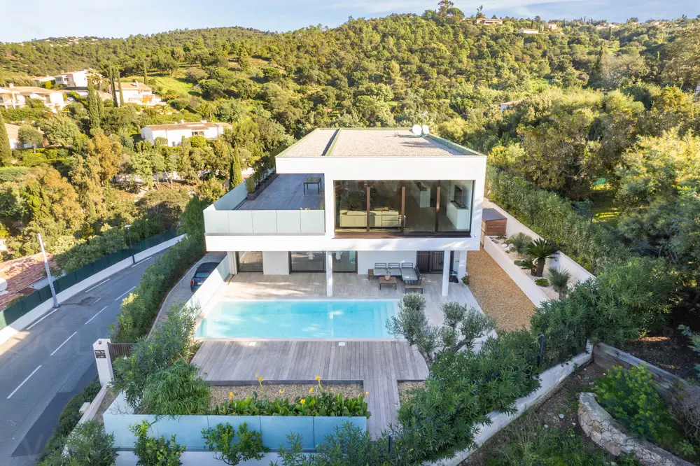 Aerial view of a modern, two-story white house with a pool and wooden deck, surrounded by lush greenery and a hillside backdrop.