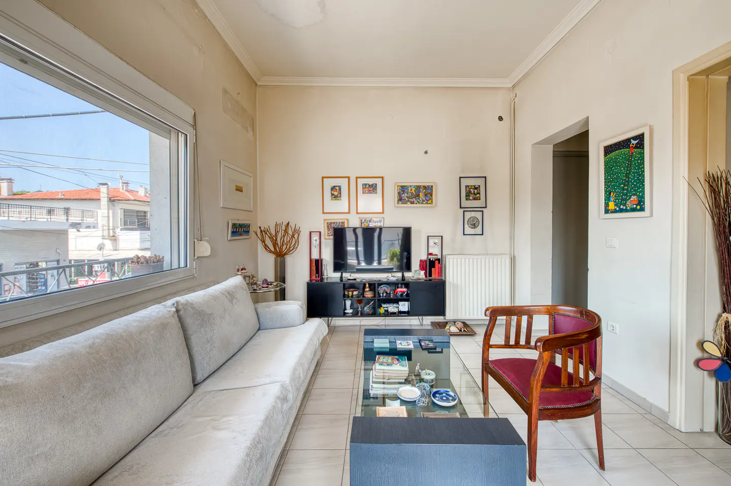 Bright living room with a grey sofa, glass coffee table, wooden chair, and a TV on a black console against a gallery wall.