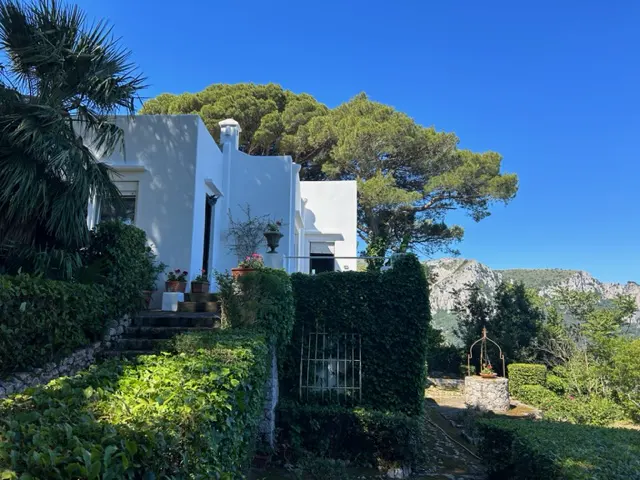 Exterior view of a white villa with lush green foliage, stone steps, and a well against a mountain backdrop under a clear blue sky.