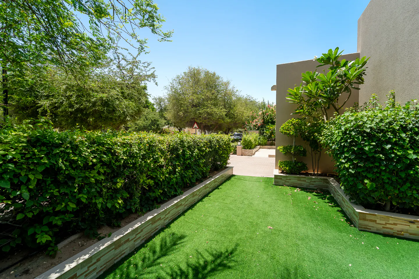 Landscaped backyard with green grass, trimmed hedges, and trees under a clear blue sky. A beige building is on the right.