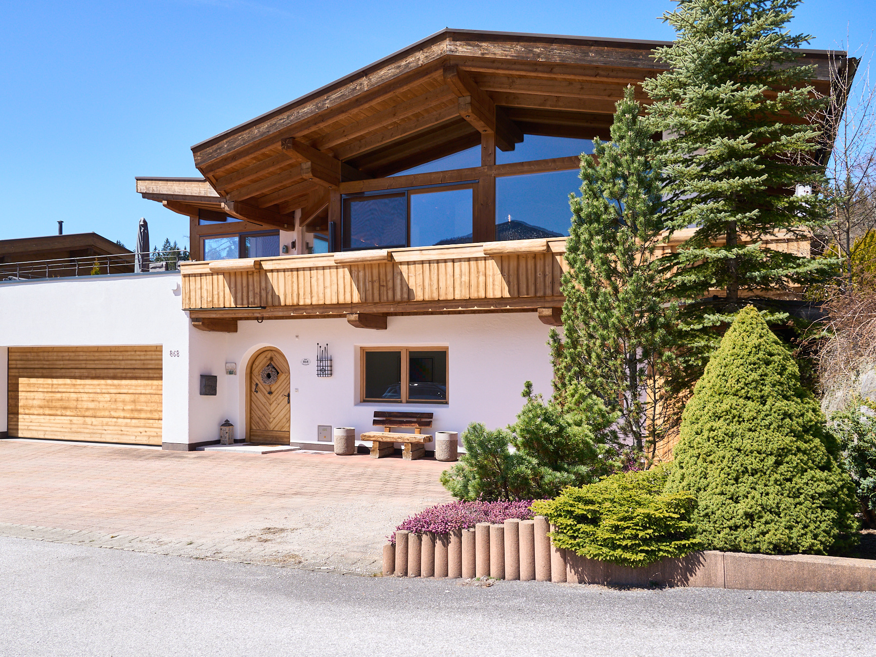Modern chalet-style home with a wooden balcony, white lower level, garage, and landscaped front yard under a blue sky.