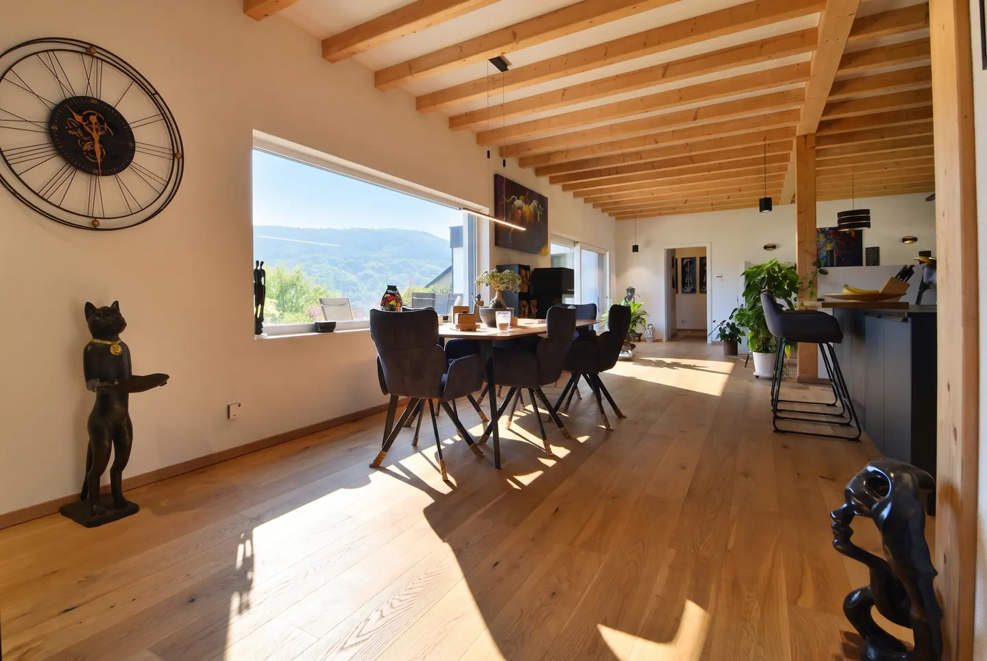 Modern dining area with wooden ceiling beams, large window with mountain view, dark chairs, and decorative cat statues.