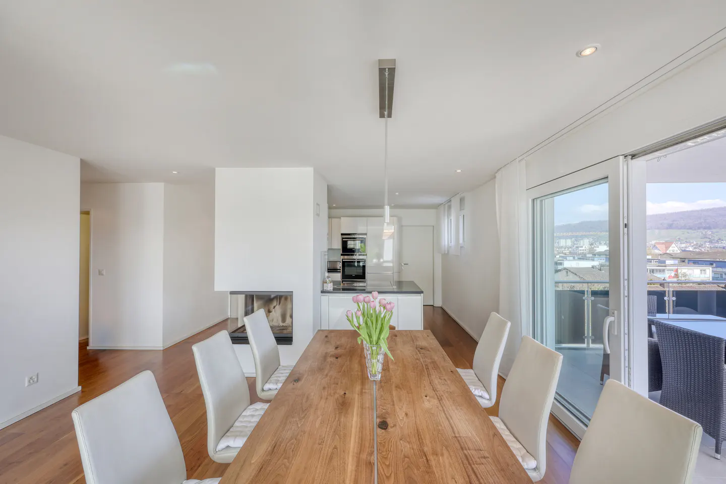 Bright, modern dining room with a long wooden table, white chairs, and a vase of pink tulips. A fireplace and kitchen are in the background. Sliding glass doors lead to a balcony with a view.