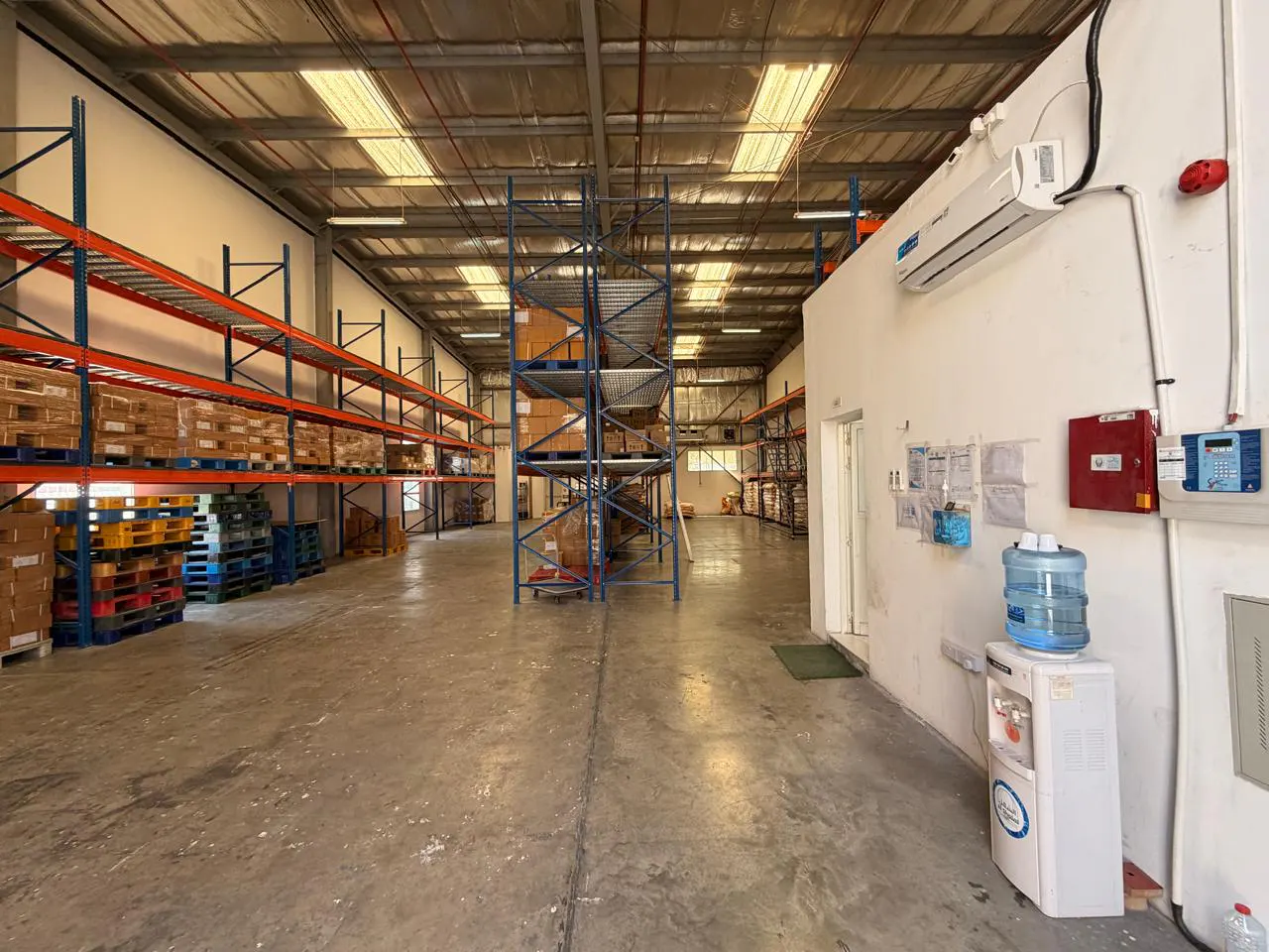 A wide shot of a warehouse interior with metal shelving, pallets, and a water cooler.
