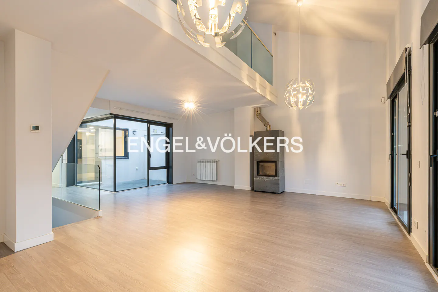 Bright, modern living room with wood floors, white walls, and a stone fireplace. Chandeliers hang from the high ceiling. Glass doors lead to a patio.