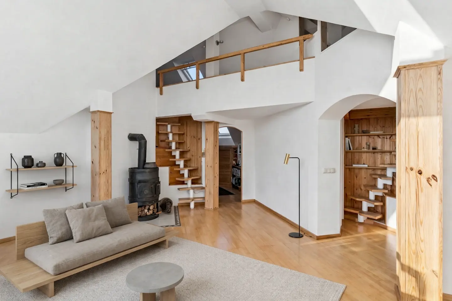 Bright living room with wood floors, white walls, and a loft. A wood-burning stove sits near a modern sofa and open staircase.