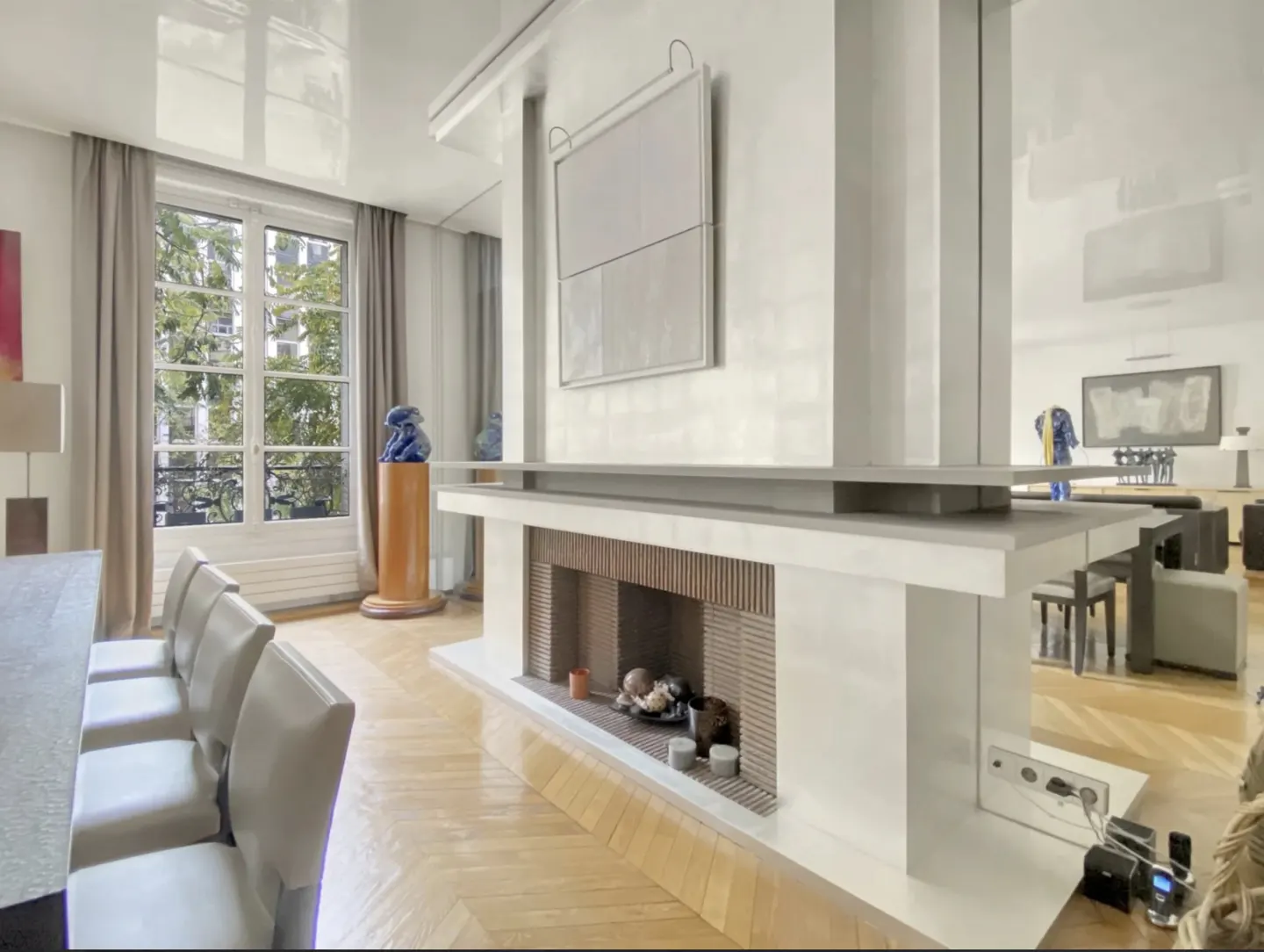 Bright living room with herringbone wood floors, a white fireplace, and a window with a view of trees.