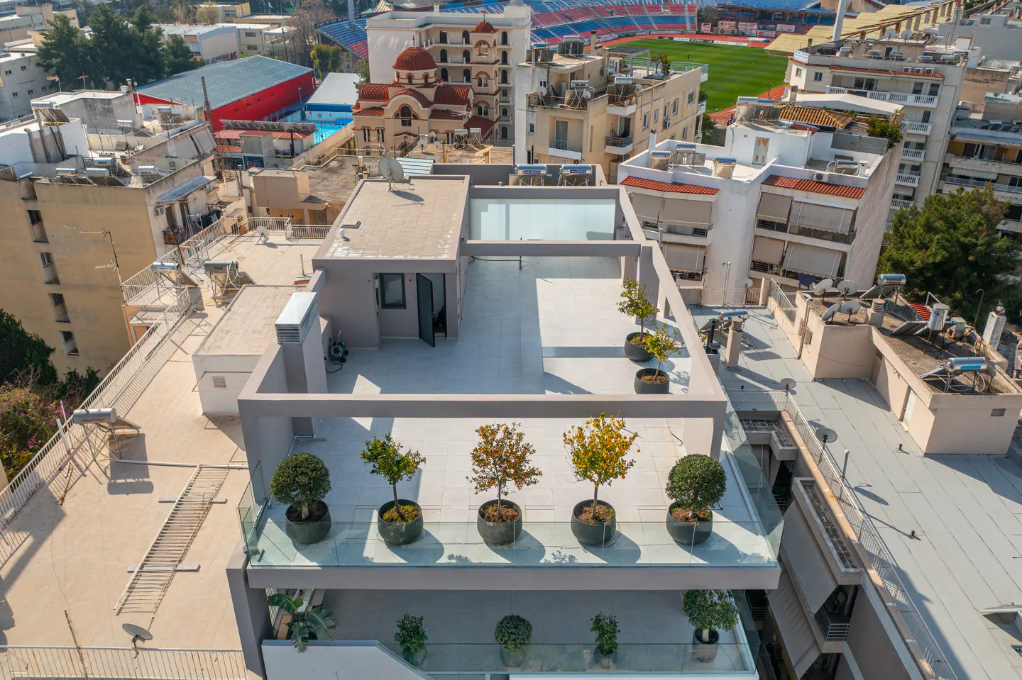 Aerial view of a modern rooftop terrace with potted trees, glass railings, and surrounding city buildings and a stadium.