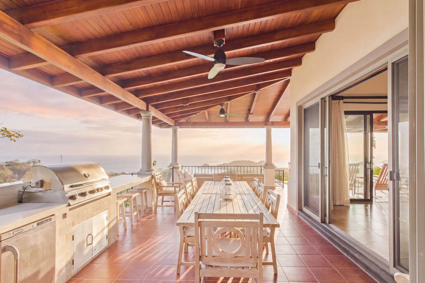 Outdoor patio with a long wooden table, chairs, grill, and ocean view under a wood-covered ceiling with a fan.