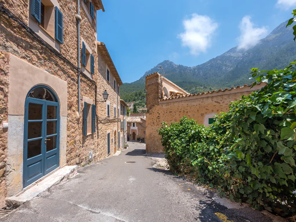 A narrow street in a European village with stone buildings and blue shutters, leading to mountains in the background.
