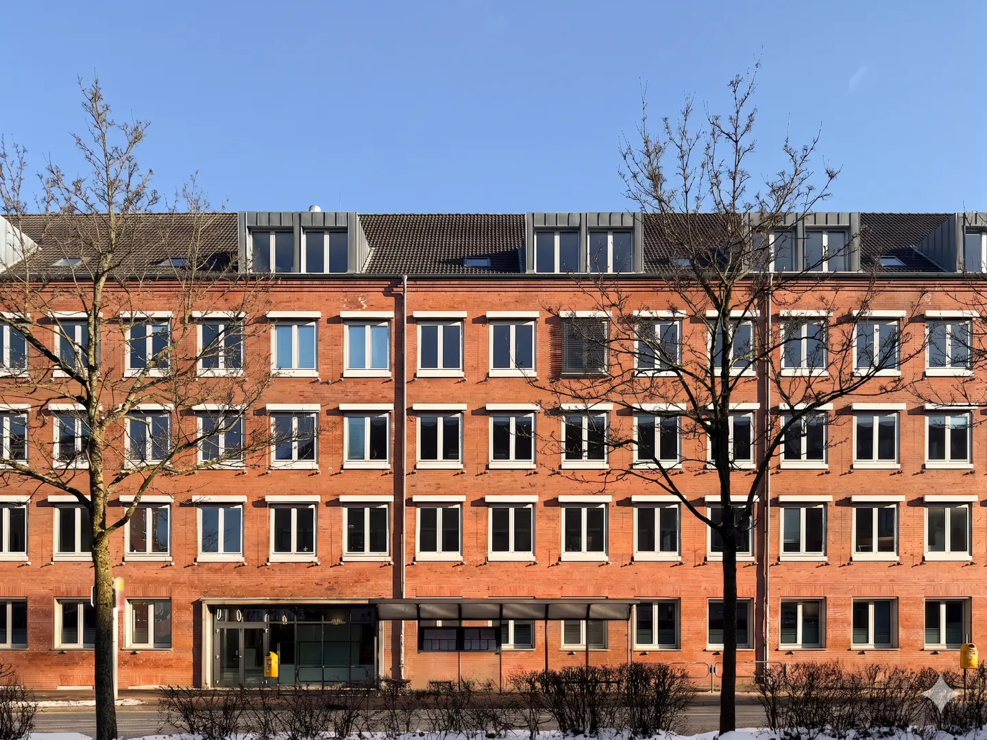 A three-story red brick building with many windows under a blue sky, framed by bare trees.