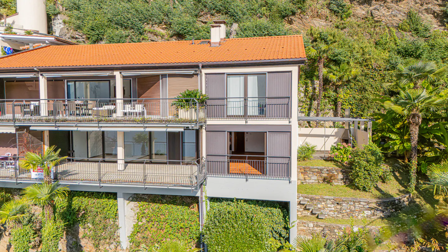 Exterior view of a three-story building with balconies, an orange roof, and lush greenery.