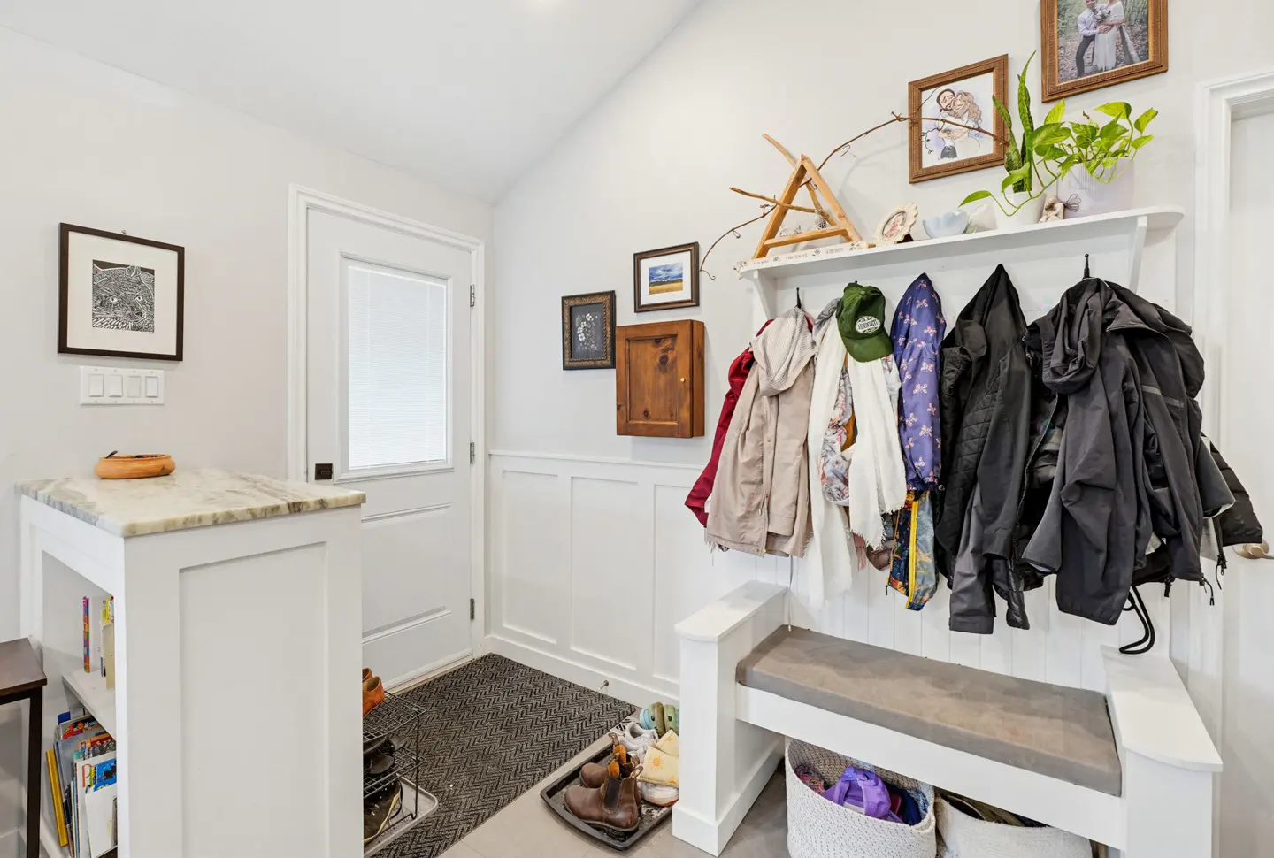 Bright entryway with a white door, a coat rack bench with hanging jackets, a shoe tray, and a small marble-topped console.