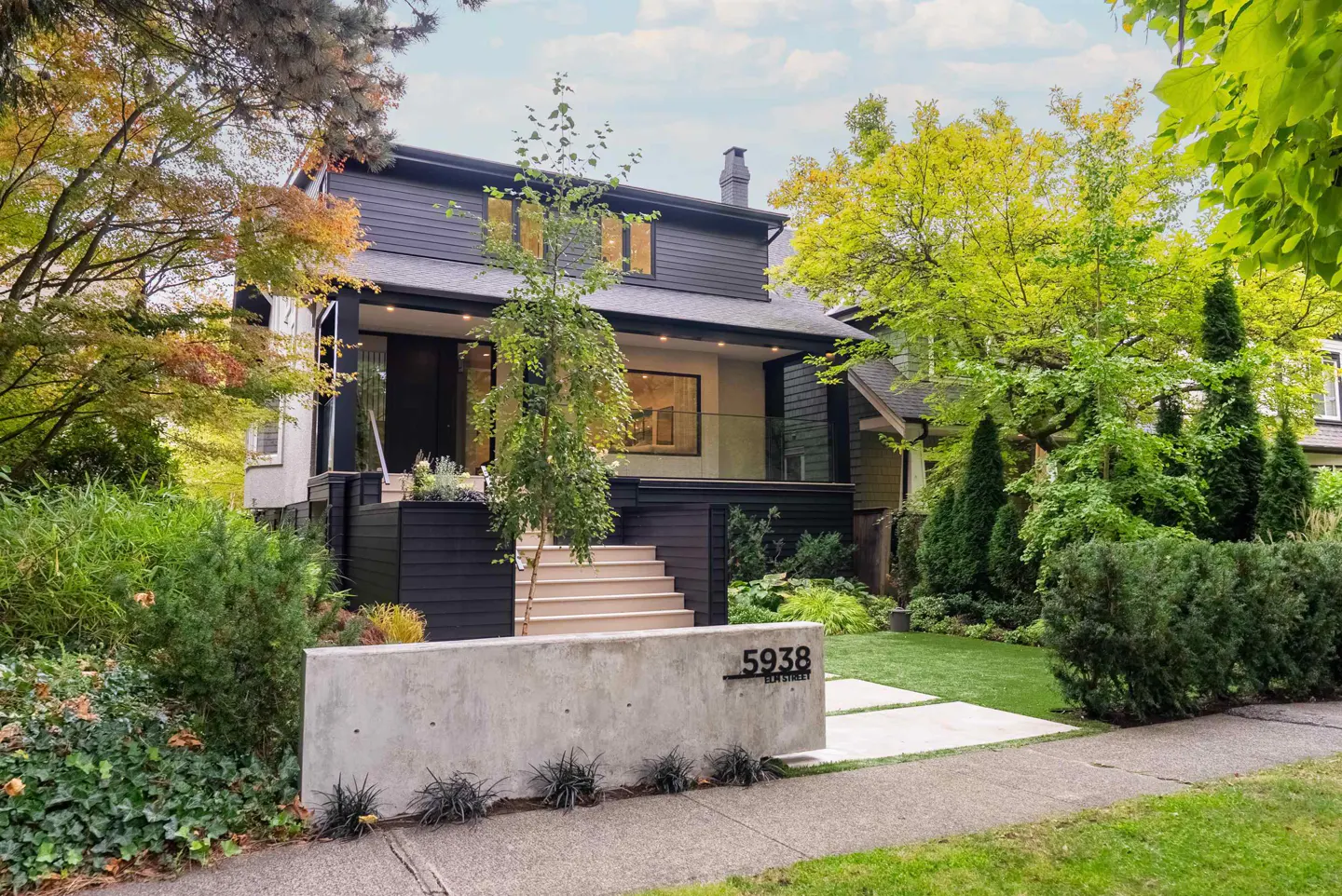 Two-story house with black siding and a concrete address sign reading "5938 Elm Street" in front. Green lawn and trees surround the property.