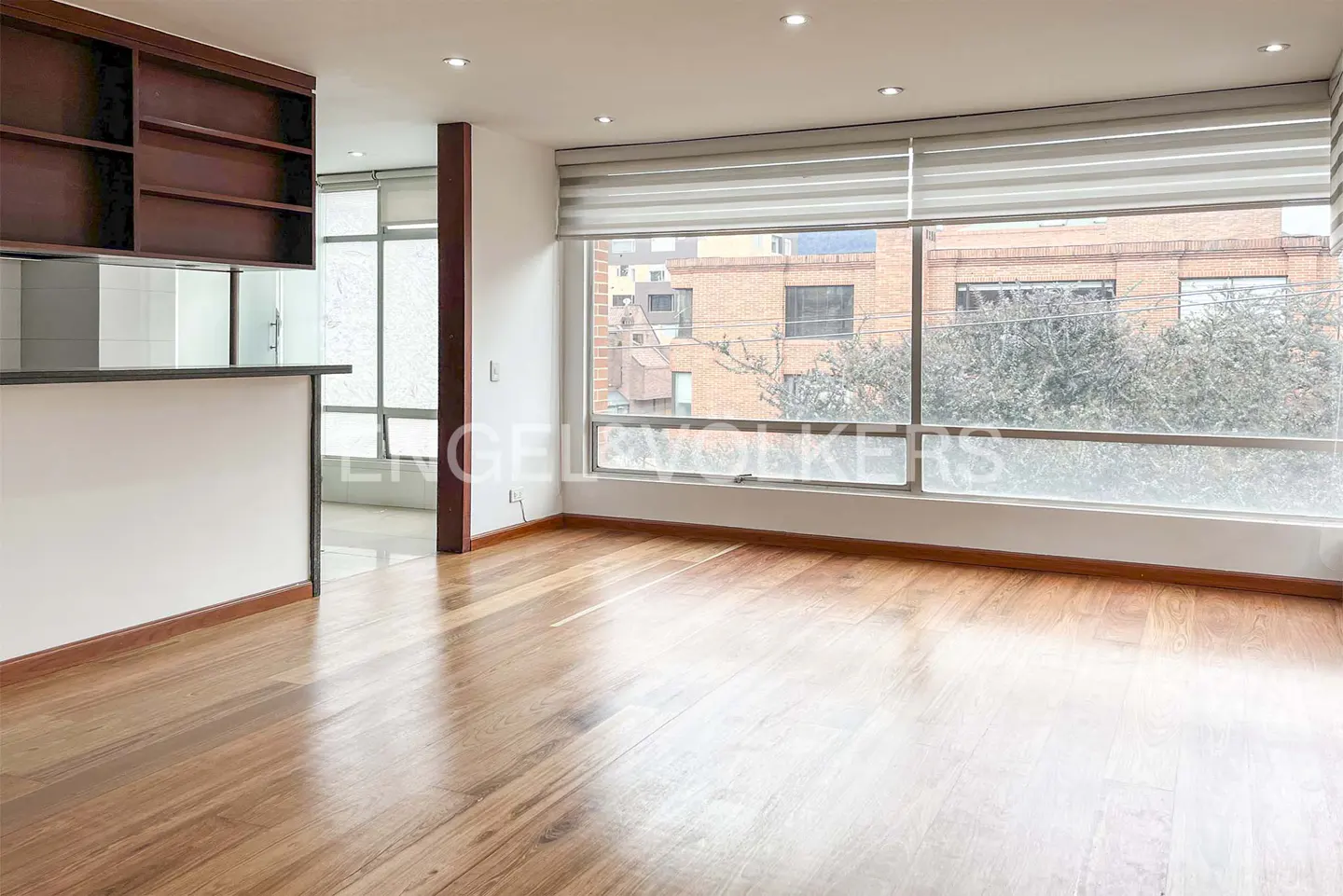 Bright, empty room with wood floors, large windows with blinds, and a dark wood bookshelf.