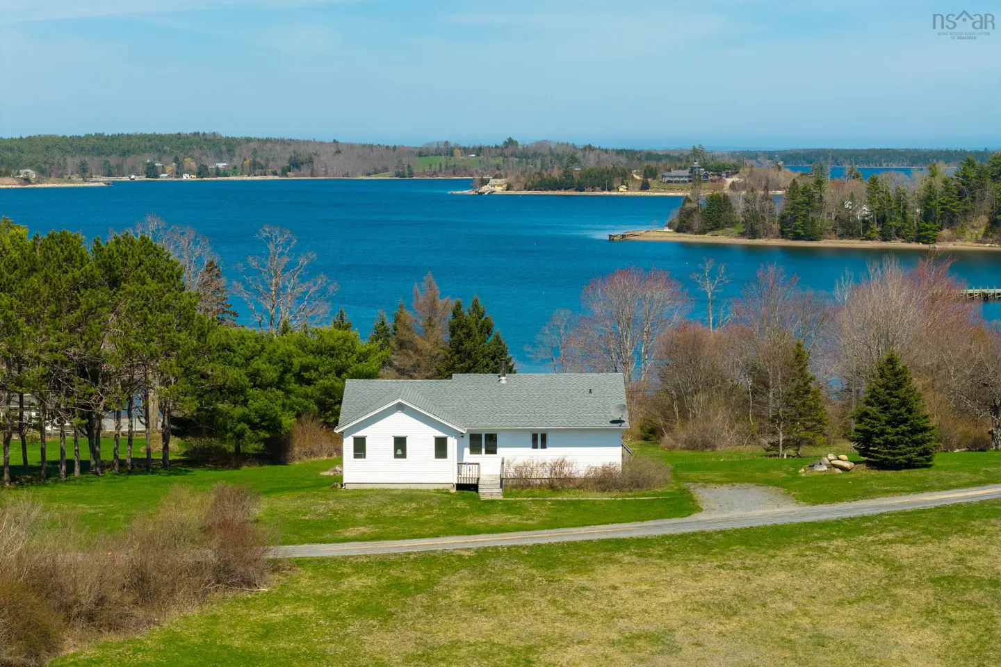 A white house with a gray roof sits on a green lawn overlooking a blue bay with wooded islands.