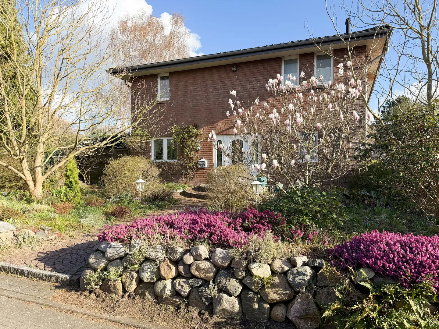 Two-story brick house with a dark roof, surrounded by lush landscaping and a stone wall with purple flowers.
