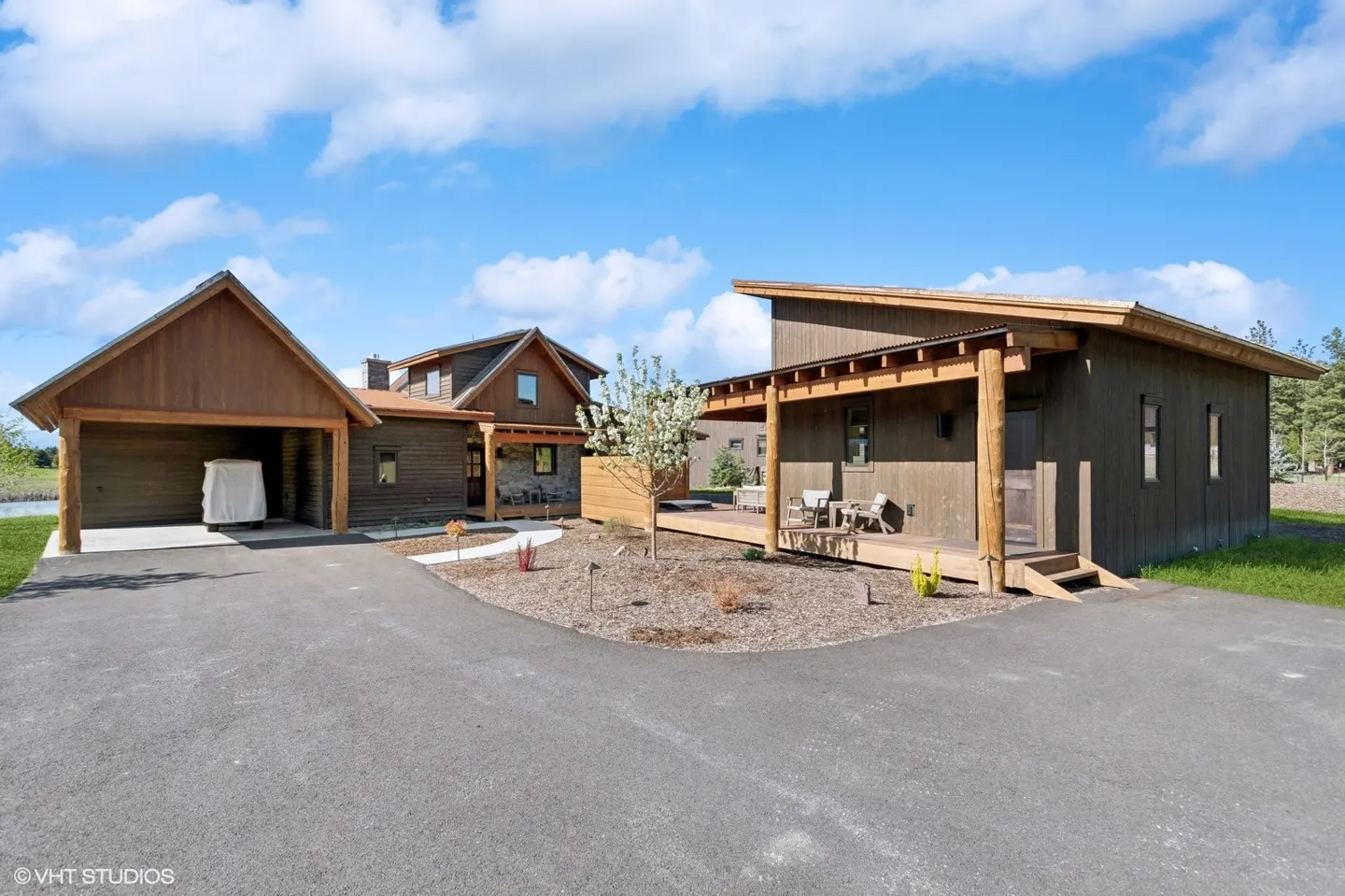 Exterior view of a modern brown house with a carport, patio, and a driveway under a blue sky.