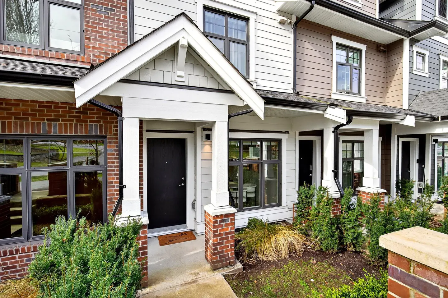 Exterior view of a modern townhouse with a black front door, white porch, brick accents, and green landscaping.