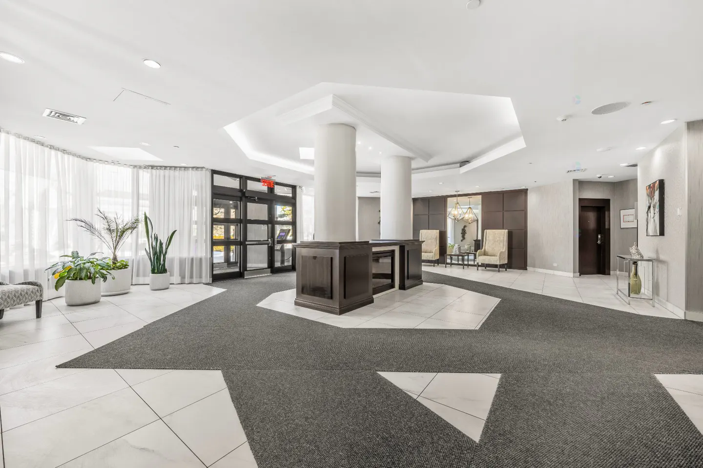 Bright lobby with white tile floors, gray carpet, and white columns. Plants near glass doors. Seating area in the background.