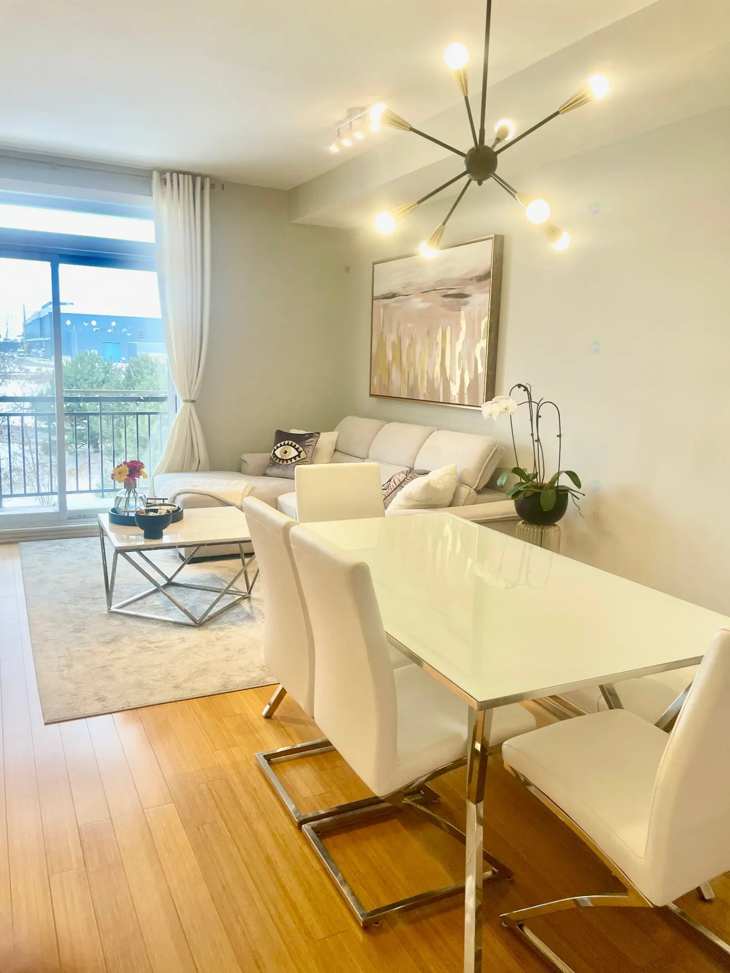 Bright, modern living and dining area with wood floors. A white sofa, table, and chairs are accented by a black chandelier and abstract art. A large window offers a view.