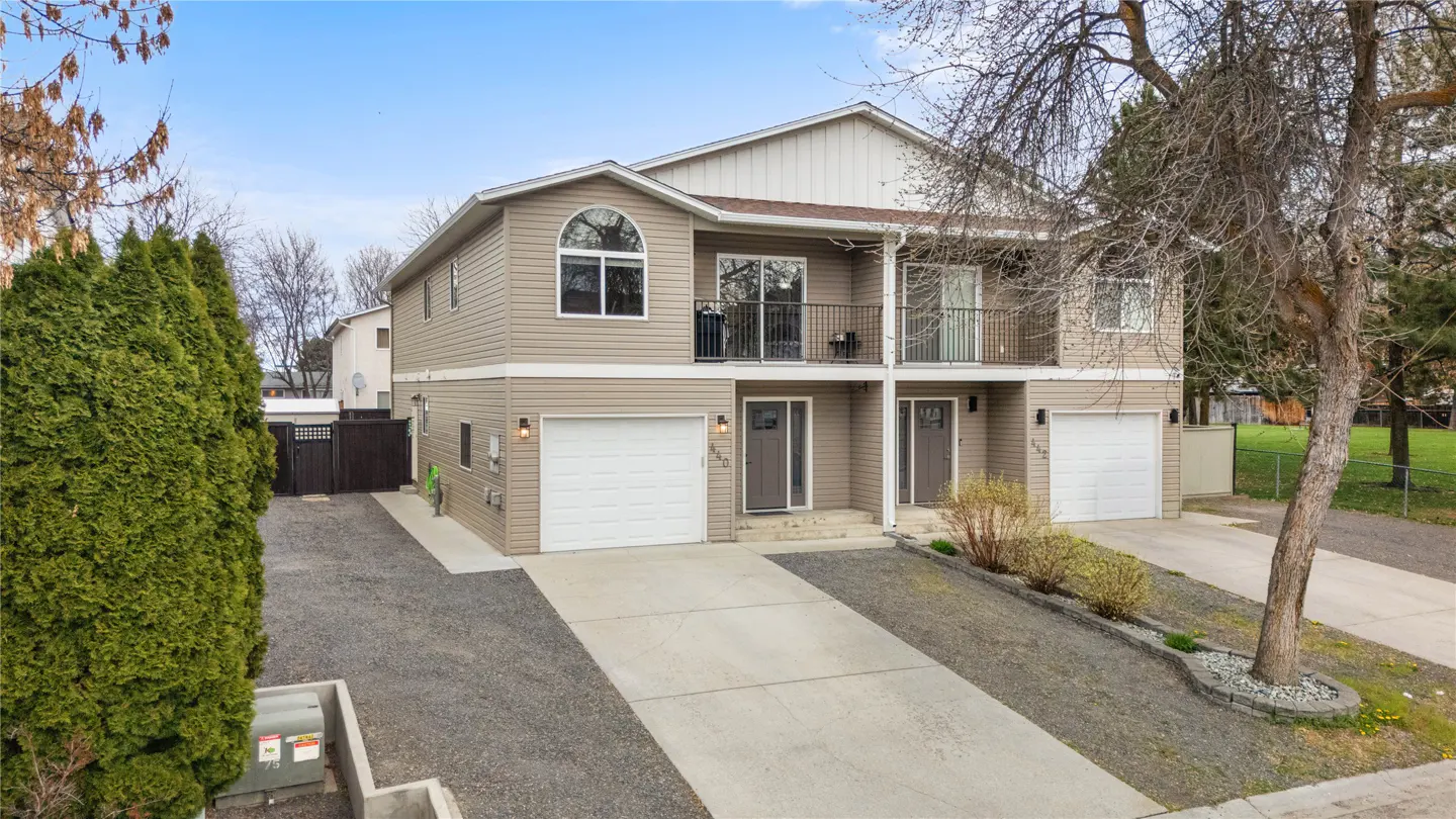 Two-story duplex with beige siding, white trim, and black iron balconies. Each unit has a garage and front door. A concrete driveway leads to the street.