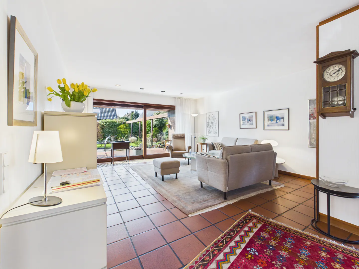 Bright living room with terracotta tile floor, beige sofa, and sliding glass doors to a green garden. A wooden clock hangs on the wall.