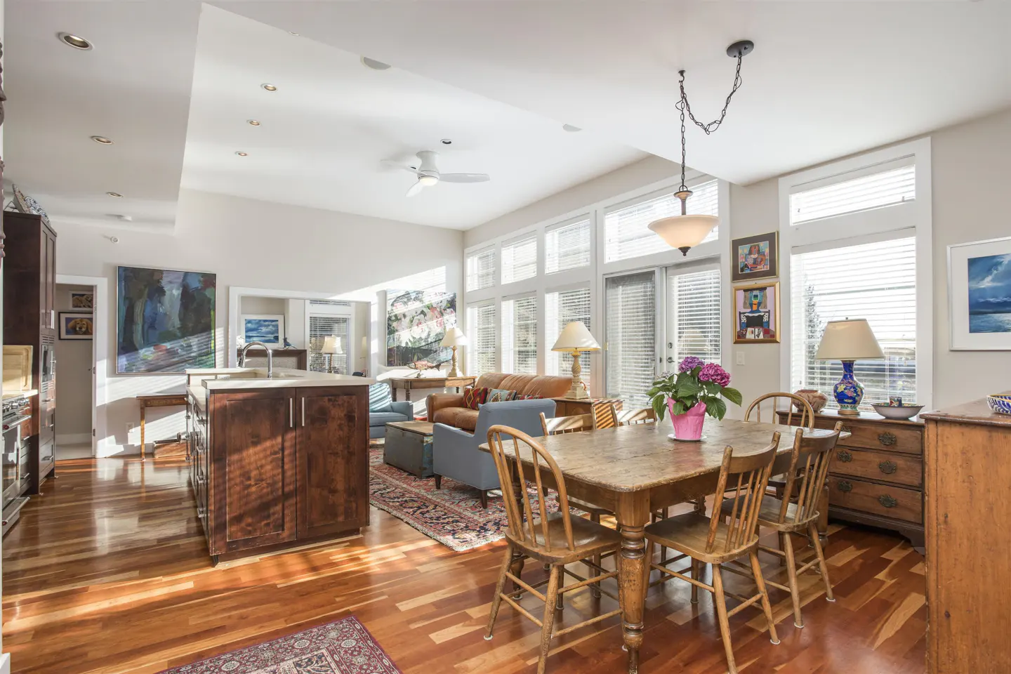 Open-concept living space with hardwood floors, dining table, kitchen island, and living room furniture. Large windows provide natural light.