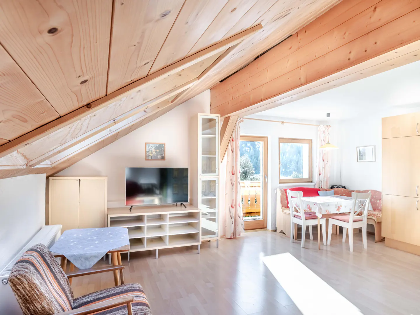 Attic apartment with wood ceiling, TV, and dining area. Balcony doors open to a view of trees.
