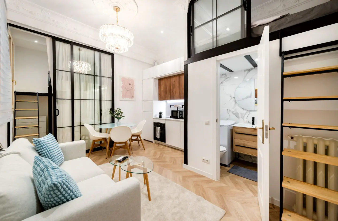 Bright, modern apartment interior with white sofa, glass table, dining area, and open bathroom. Herringbone wood floors and a chandelier add elegance.