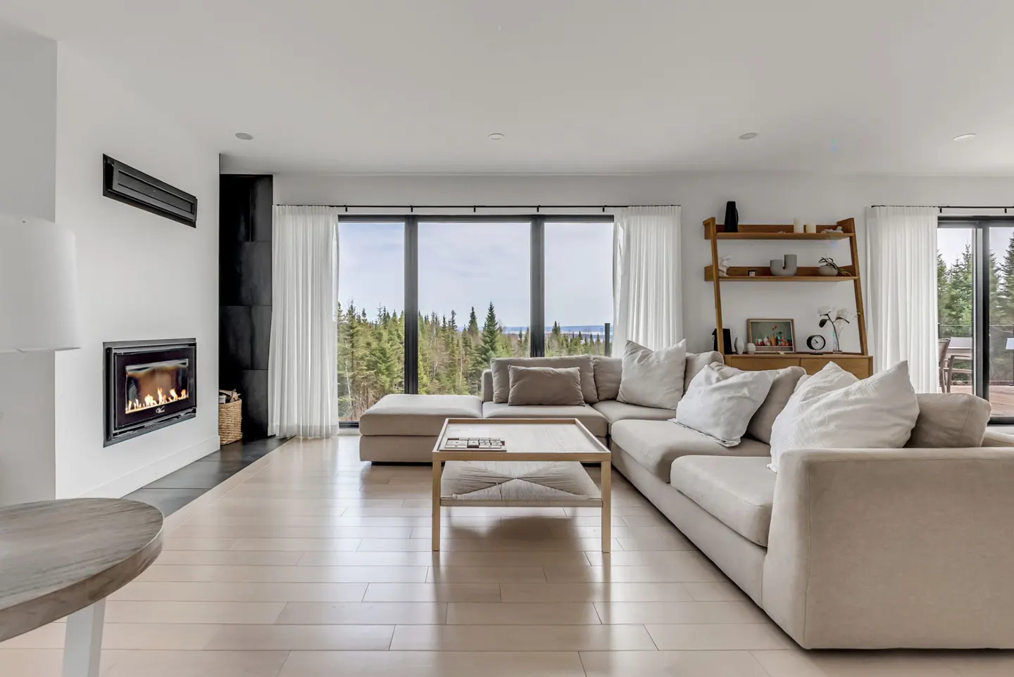 Bright living room with a beige sectional sofa, light wood floors, and a fireplace. Large windows overlook a forest.