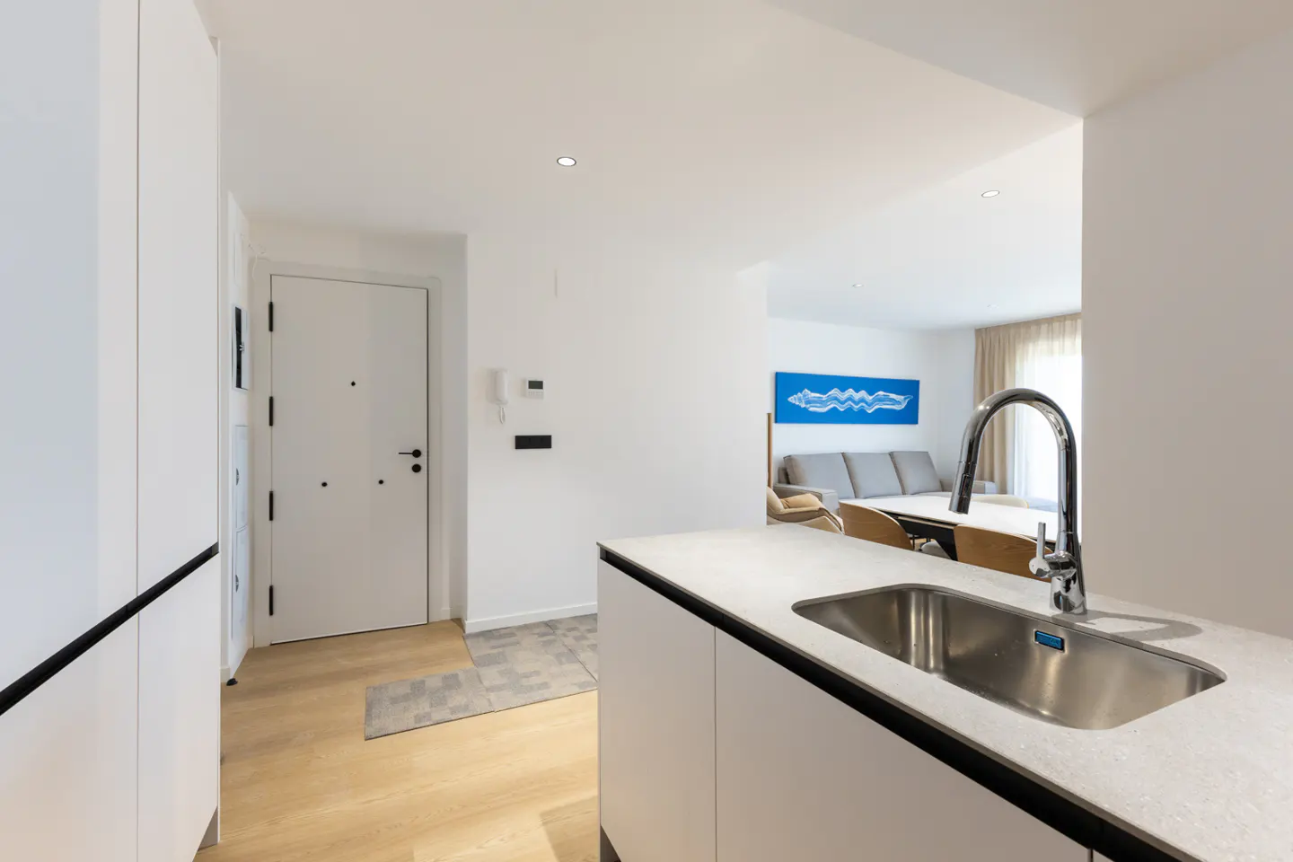 Modern apartment interior with a stainless steel sink, white cabinets, and a view into the living room with a gray sofa.