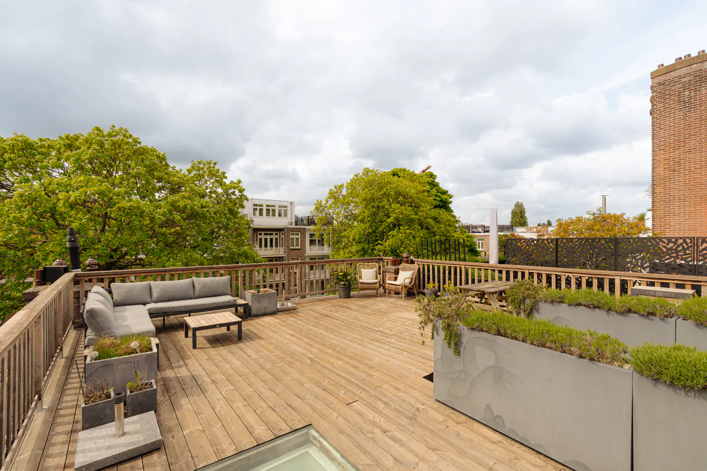 Wide shot of a wooden rooftop terrace with gray sofa, chairs, planters, and a wooden fence, with trees and buildings in the background.