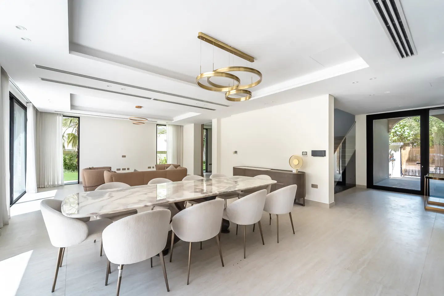 Bright, modern dining room with a marble table, white chairs, and a gold ring chandelier. A tan sofa is visible in the background.