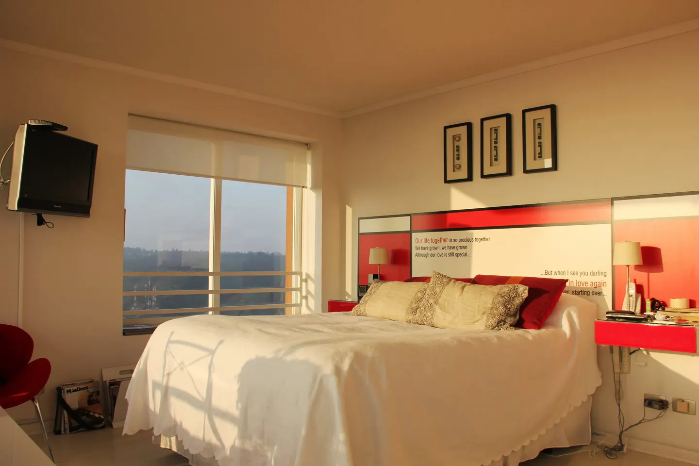 Bright bedroom with white bedding, red accents, and a large window overlooking a cityscape. A TV is mounted on the wall.