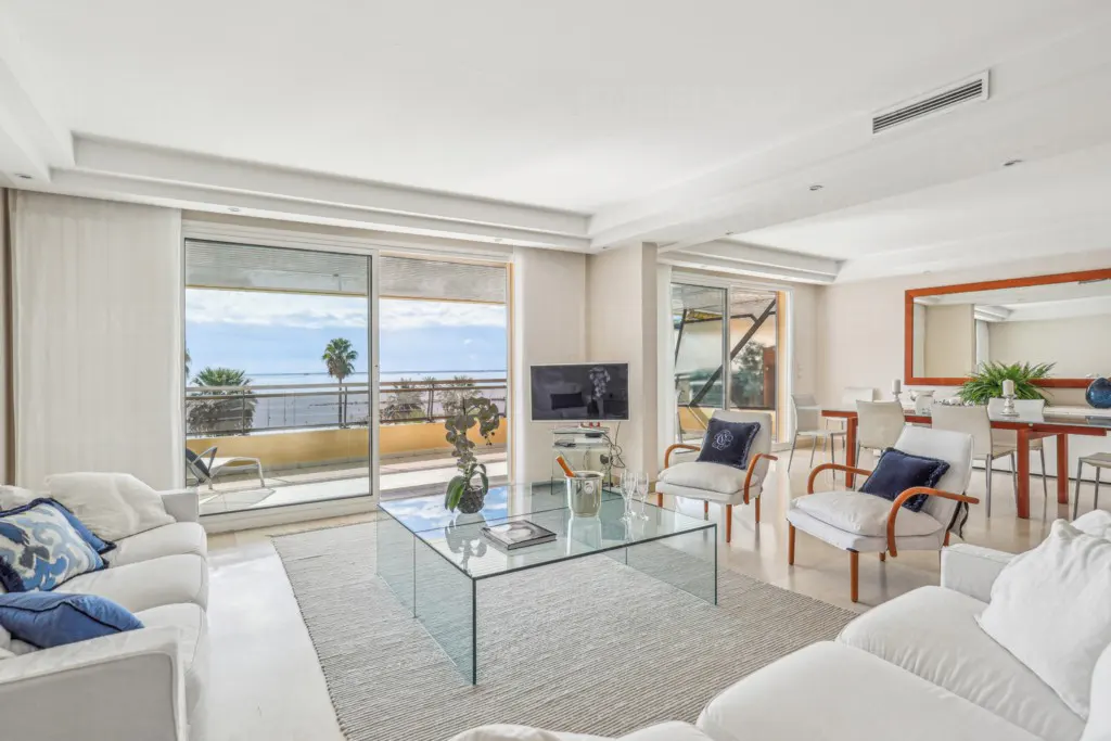 Bright living room with white sofas, chairs, and a glass table. Ocean view through sliding glass doors. Dining area visible in the background.