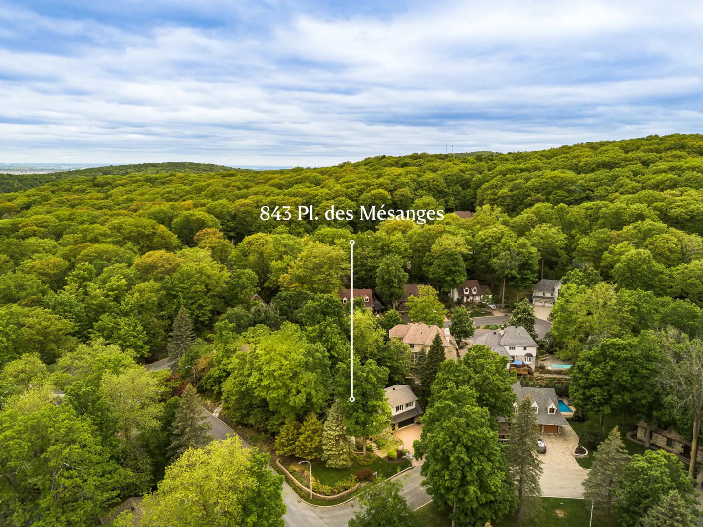 Aerial view of a house at 843 Pl. des Mésanges, nestled in a lush green forest under a cloudy sky.
