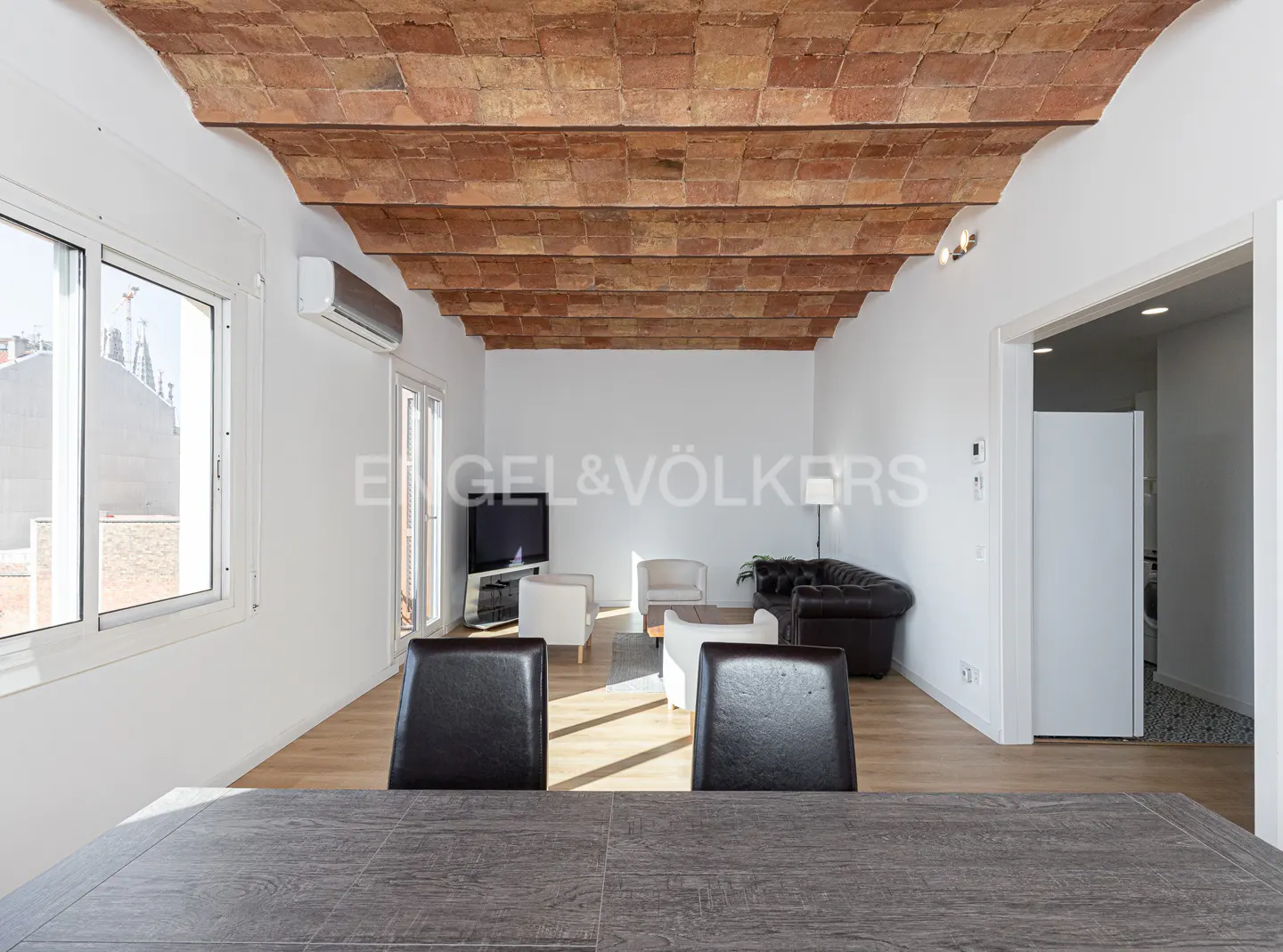 A bright living room with a brick ceiling, white walls, and wood floors. A dark table and chairs are in the foreground. A brown leather sofa is in the background.